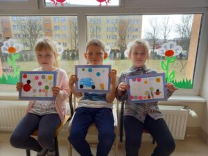 Dzieci siedząc na krzesełkach na tle okna i wiosennych dekoracji prezentują swoje prace plastyczne na temat "Kraina kół i kółeczek"