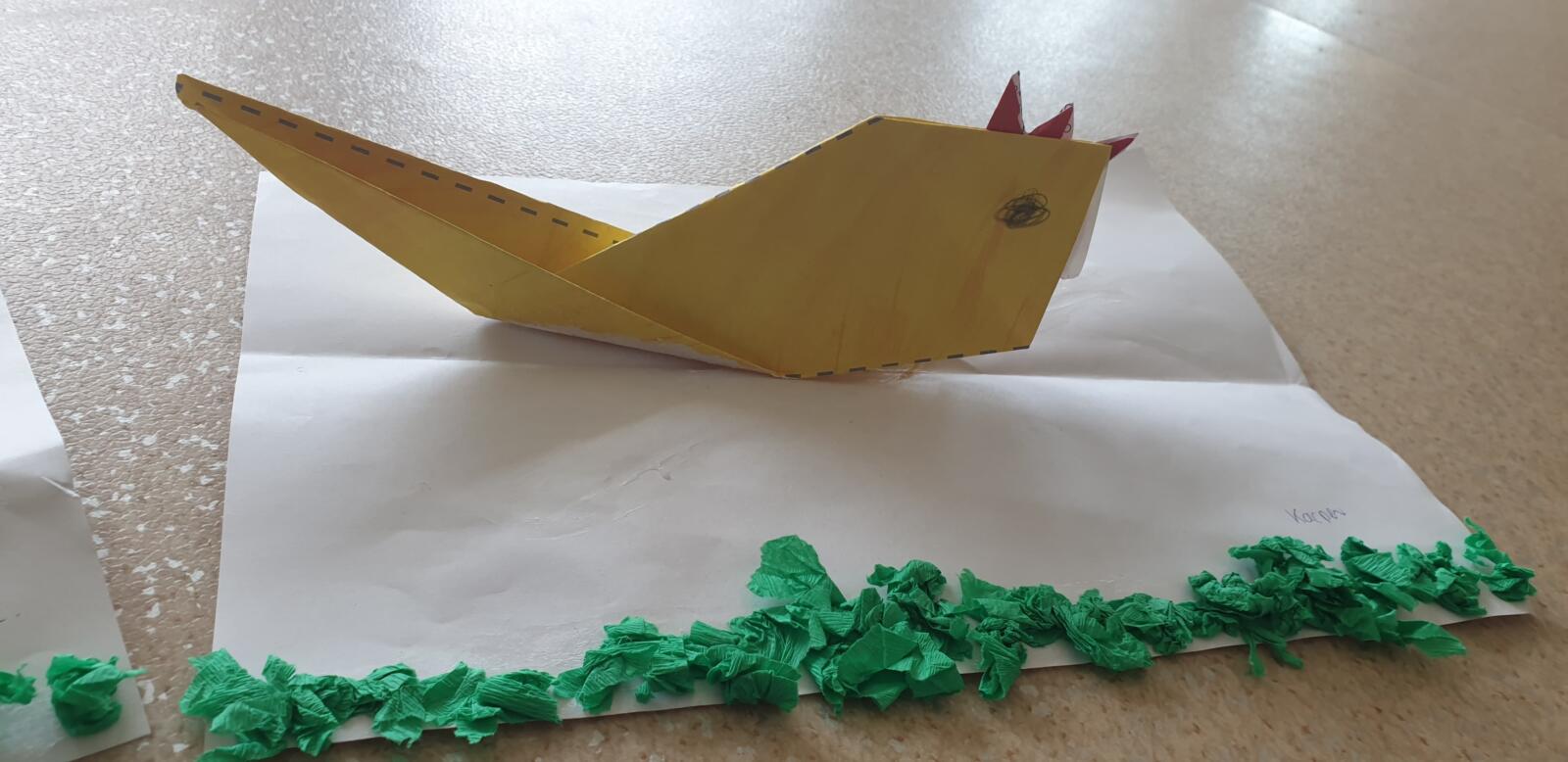 Zdjęcie przedstawia gotową pracę plastyczna jednego z przedszkolaków. Jest to żółta, papierowa kurka, przyklejona do białej kartki papieru ozdobionej zielonymi kuleczkami z krepy symbolizującej trawę.