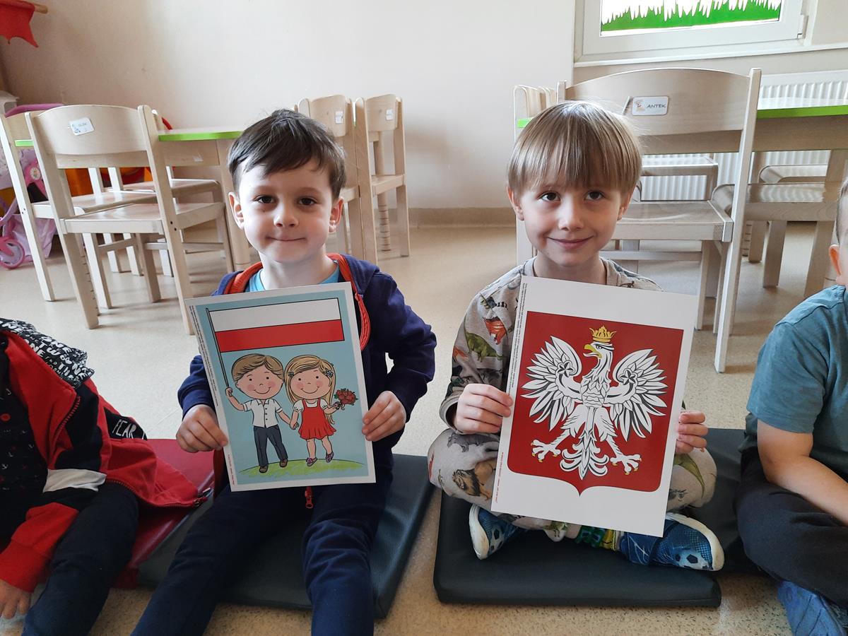 Na zdjęciu znajdują się dwaj chłopcu: pierwszy od lewej trzyma w rękach ilustrację z flagą Polski, drugi chłopiec trzyma w rękach ilustrację z godłem Polski.