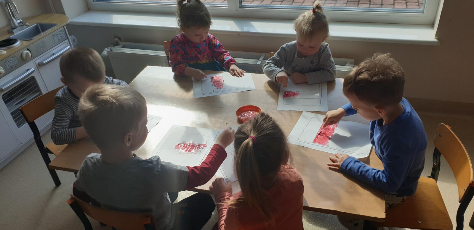 Dzieci siedzą przy stoliku i nakładają czerwoną farbkę na obrys flagi Polski. 