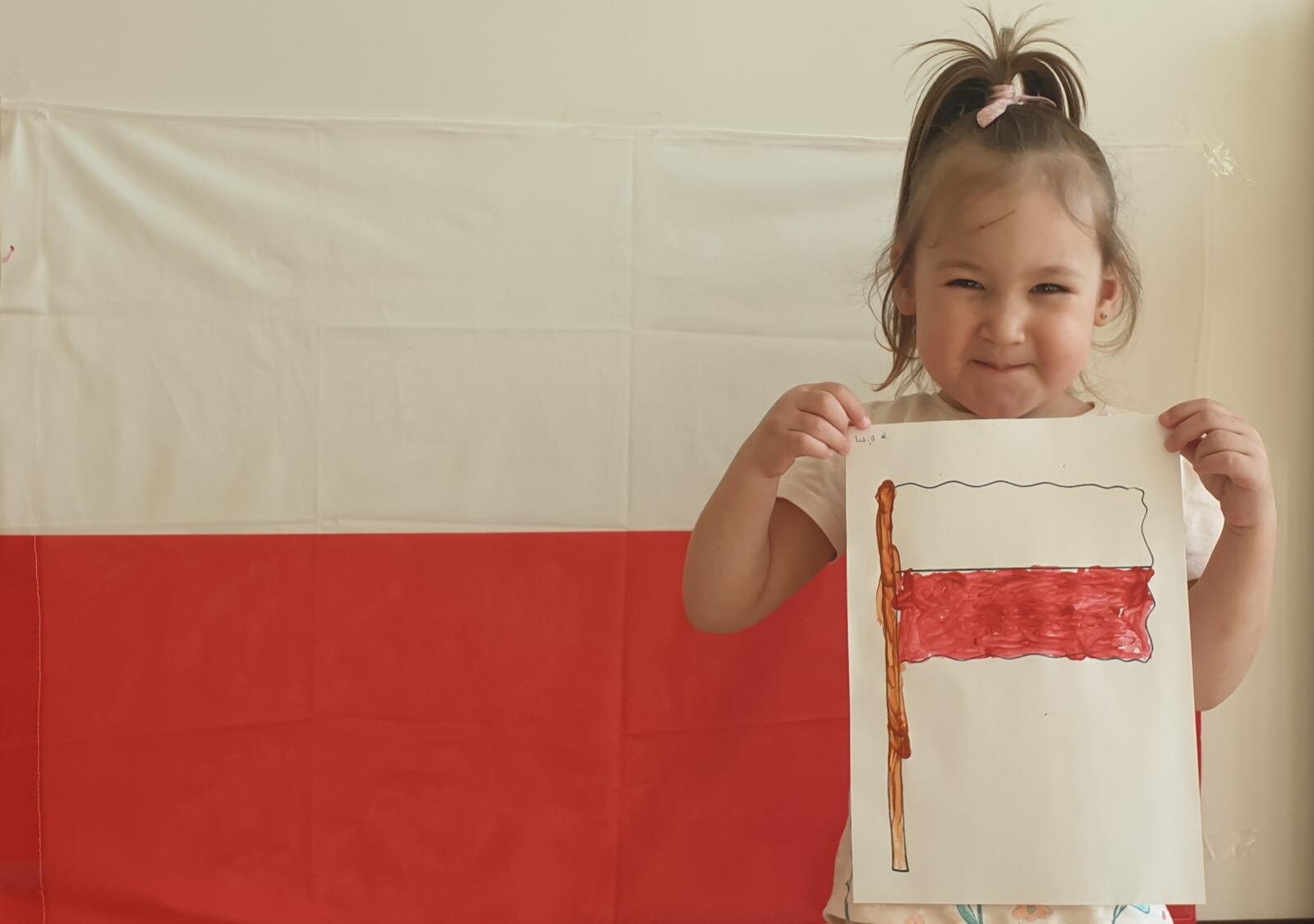 Uśmiechnięta dziewczynka pozuje ze swoją gotową, pomalowaną flagą na tle dużej flagi Polski.