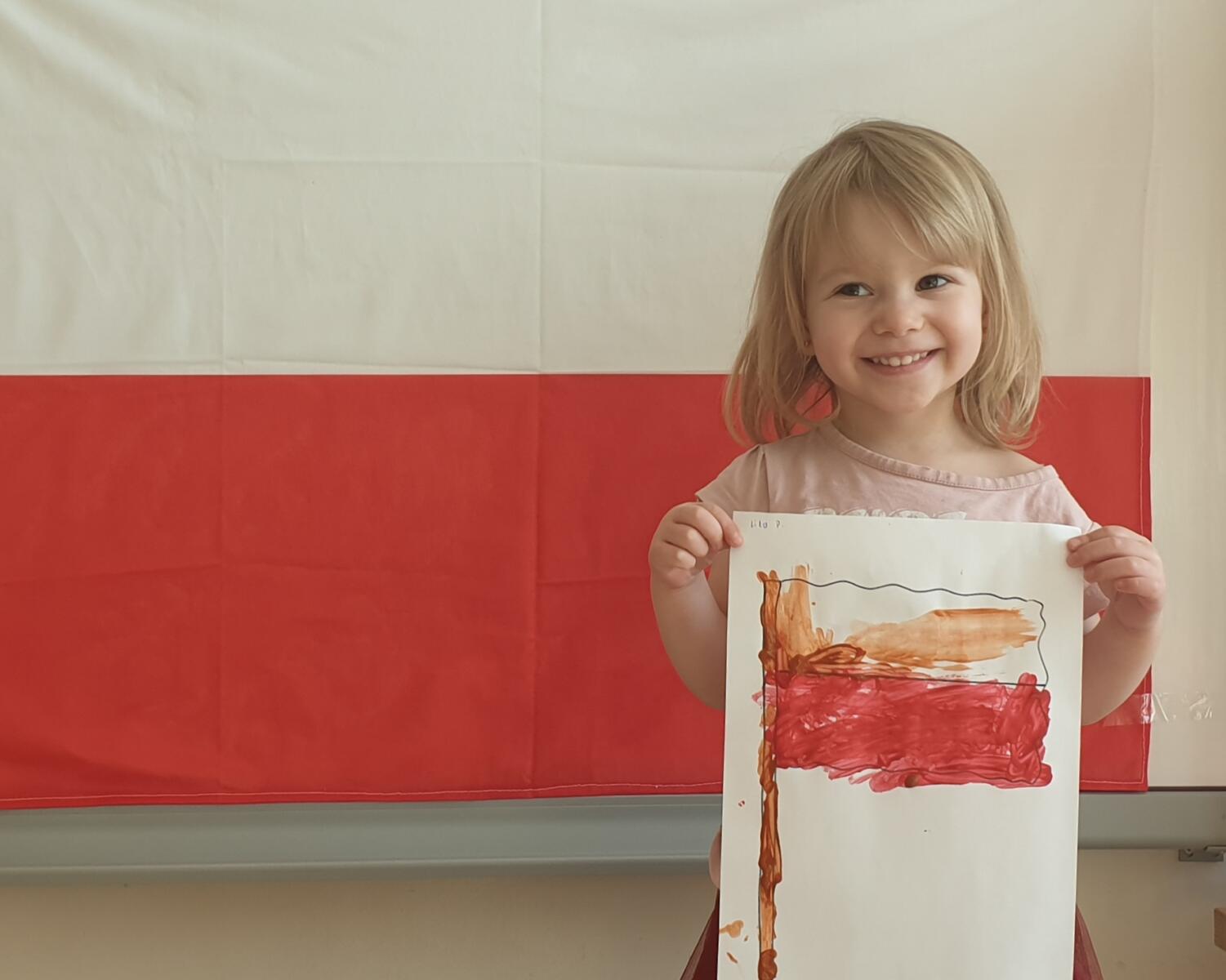 Zdjęcie przedstawia zadowoloną dziewczynkę, która trzyma swoją pracę plastyczną i pozuje do zdjęcia na tle flagi Polski.