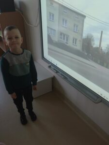 Zdjęcie przedstawia chłopca, który stoi przy tablicy multimedialnej na której wyświetla się zdjęcie jego domu. 