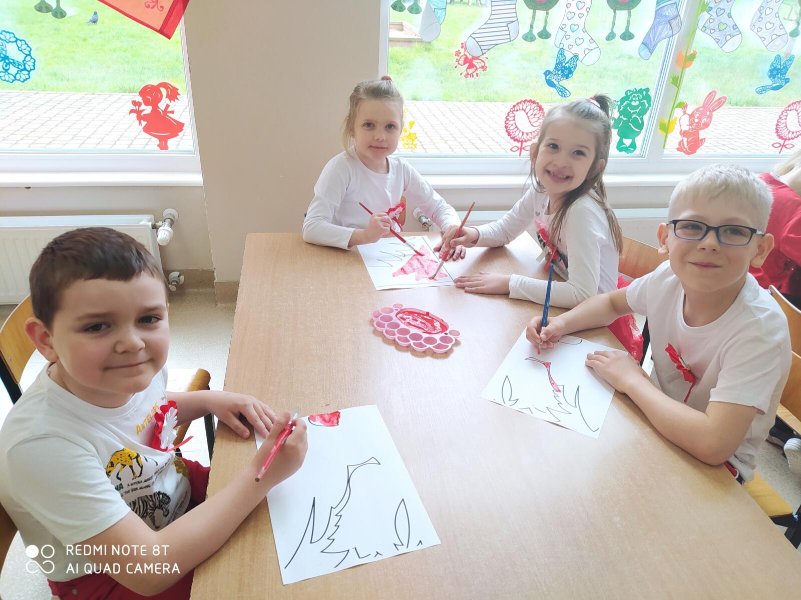 Zdjęcie przedstawia czwororo dzieci, które pracują przy stolikach w parach. Dzieci zamalowują odpowiednie pole na kartce na czerwono do wspólnej grupowej pracy - godła Polski.