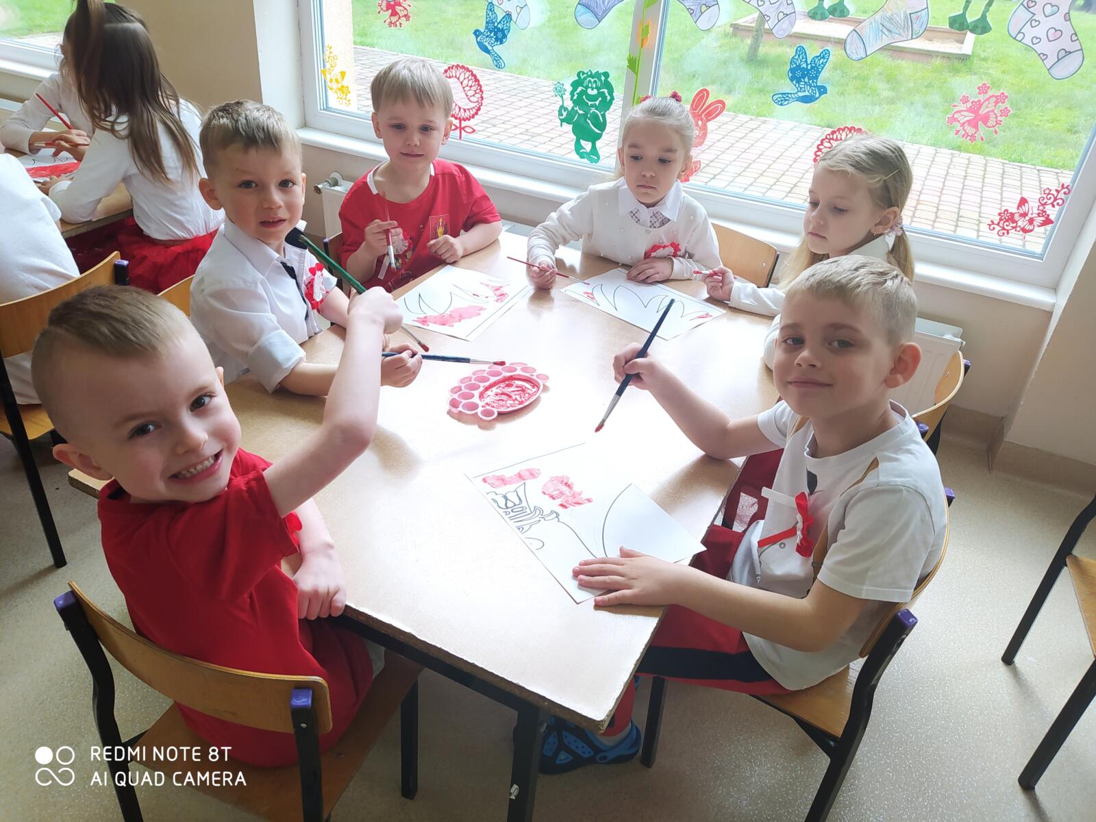 Zdjęcie przedstawia sześcioro dzieci, które pracują przy stolikach w parach. Dzieci zamalowują odpowiednie pole na kartce na czerwono do wspólnej grupowej pracy - godła Polski.
