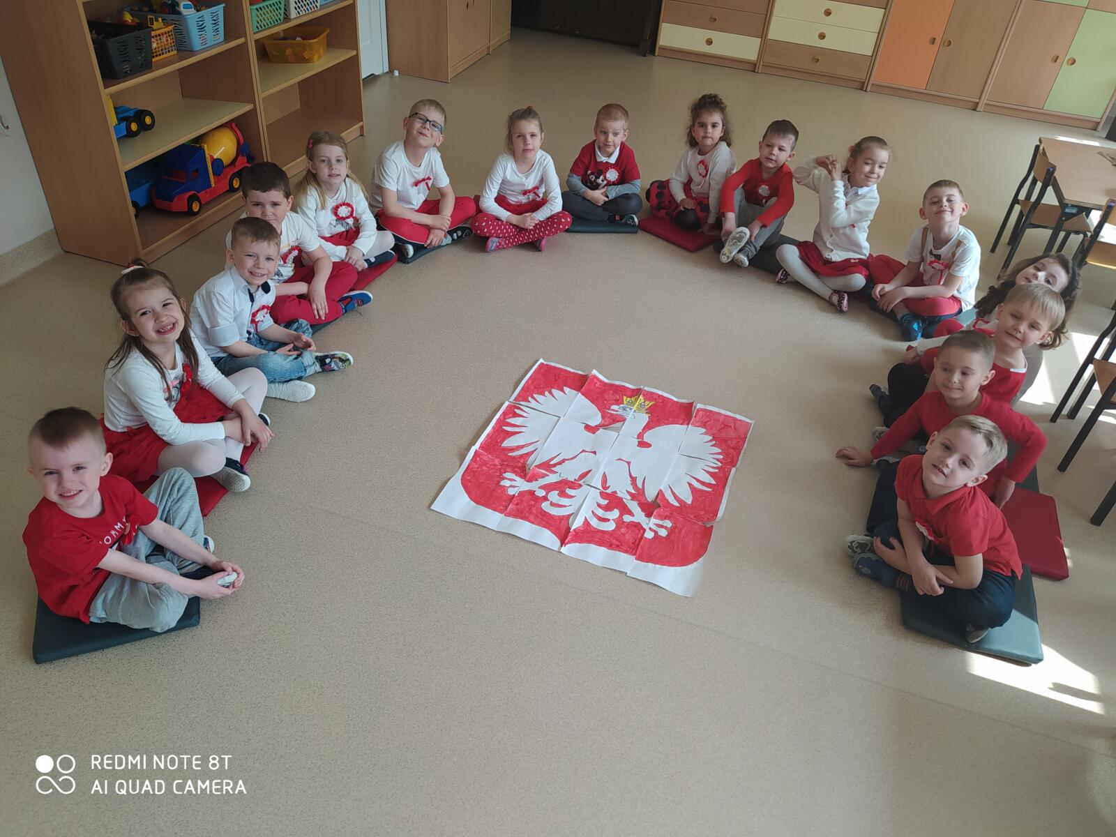 Zdjęcie przedstawia siedzące dzieci wokół godła Polski - pracy wykonanej wspólnie.