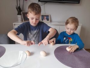 Dwóch chłopców siedzi przy stole. Chłopcy obracając jajka wykonują doświadczenia, które jajko jest surowe, które gotowane.