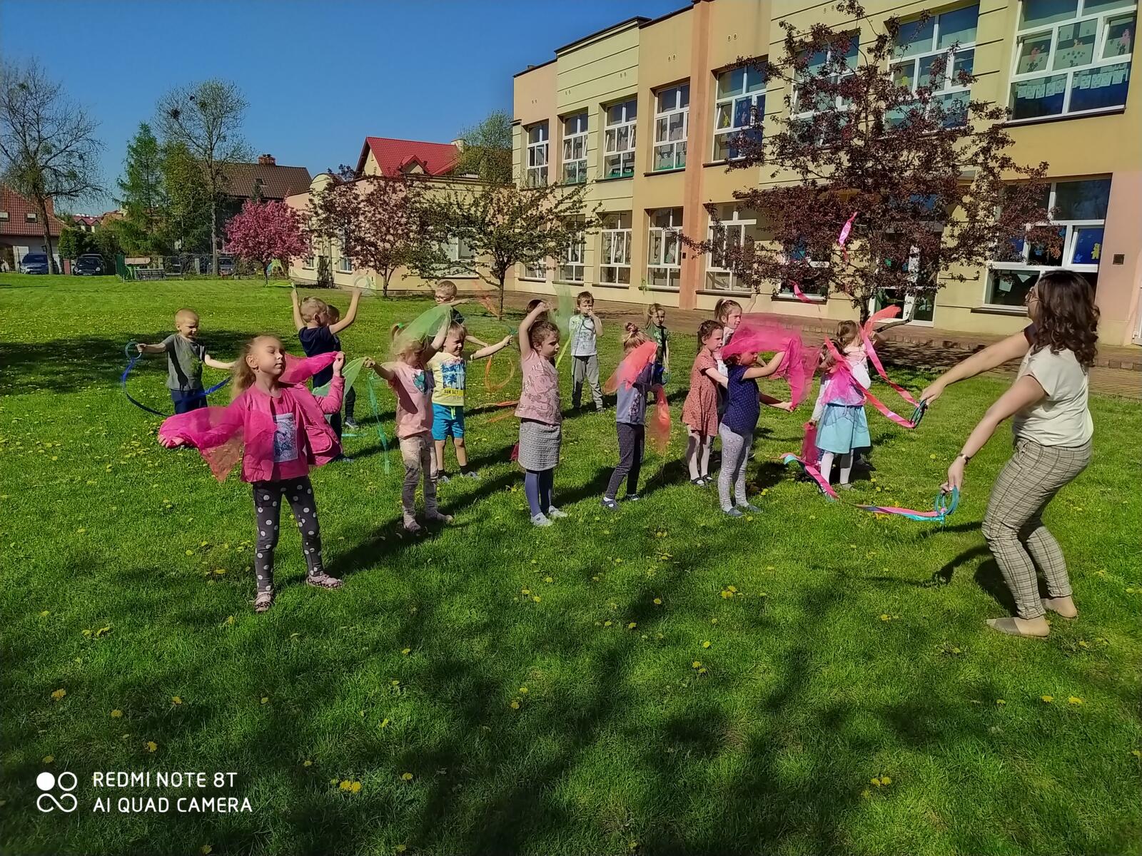 Zdjęcie przedstawia tańczące dzieci w ogrodzie przedszkolnym taniec motyli z wykorzystaniem kolorowych wstążek i chustek.