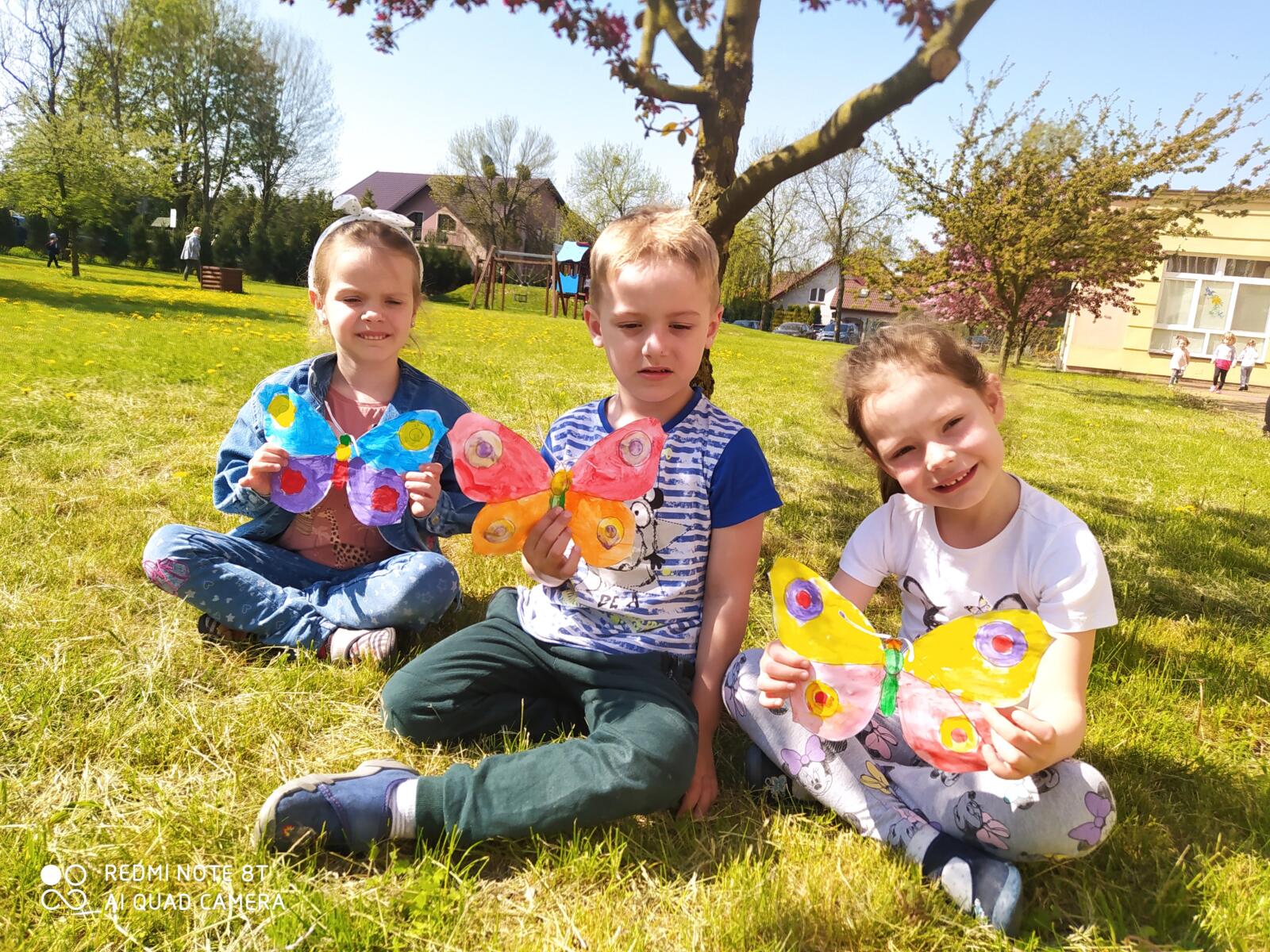 Zdjęcie przedstawia troje dzieci w ogrodzie przedszkolnym, które prezentują wykonane farbami i wycięte kolorowe motyle.