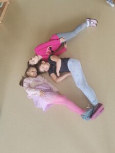 Zdjęcie przedstawia trzy dziewczynki, które leżąc na podłodze układają ze swoich ciał cyfrę 6.