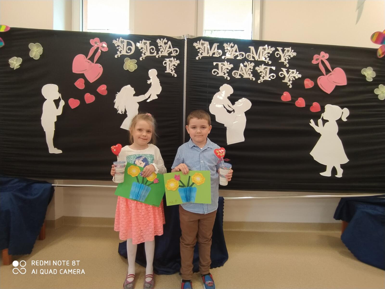 Zdjęcie przedstawia dwoje dzieci na tle dekoracji, trzymające w ręku laurki.