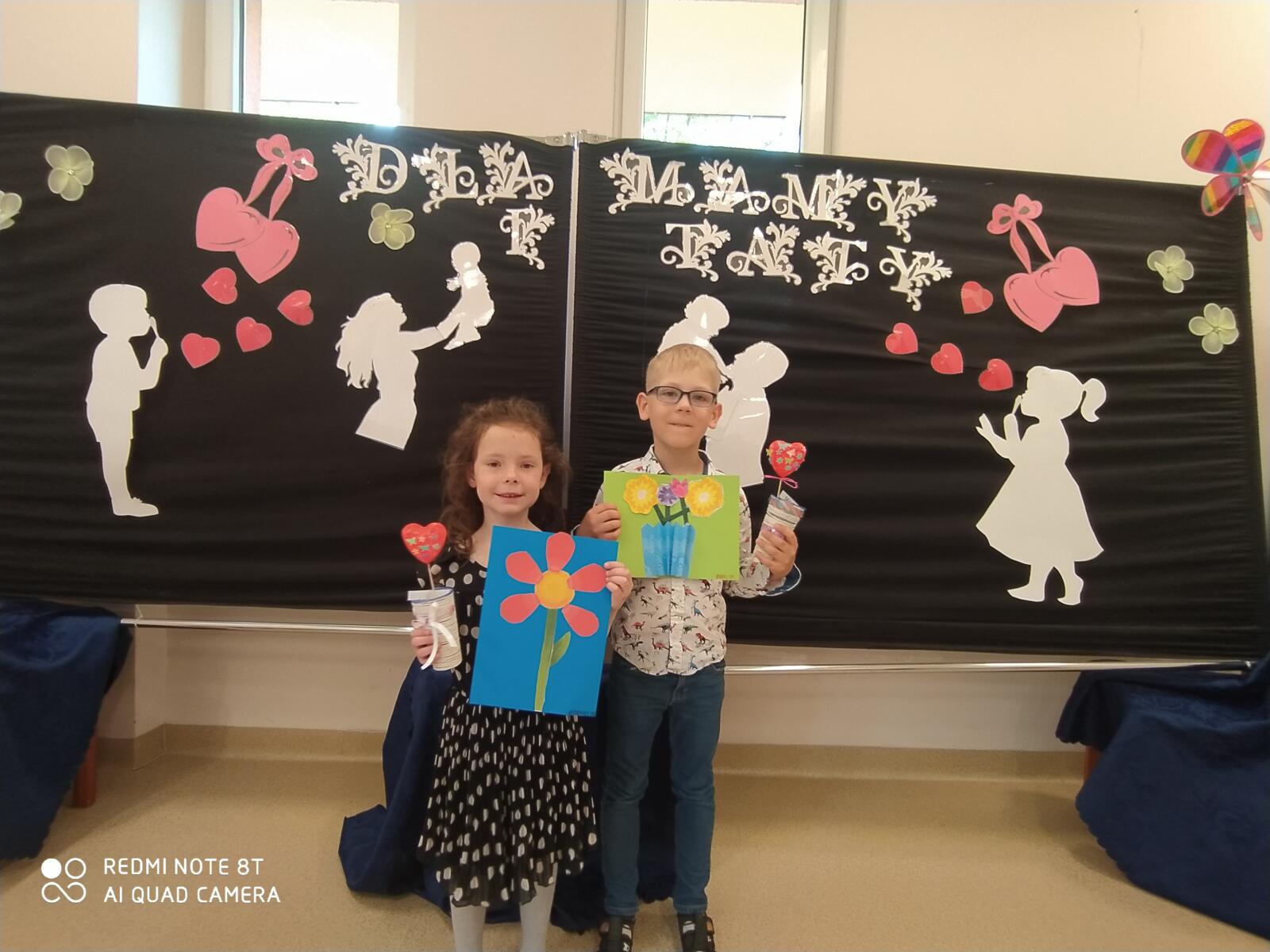 Zdjęcie przedstawia dwoje dzieci na tle dekoracji, trzymające w ręku laurki.