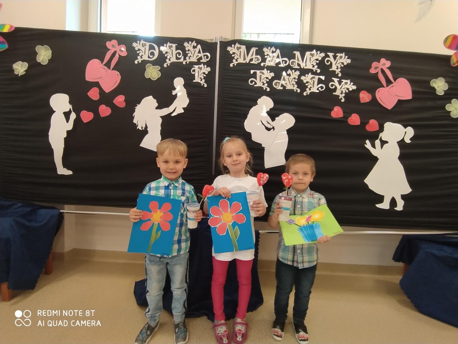 Zdjęcie przedstawia troje dzieci na tle dekoracji, trzymające w ręku laurki.