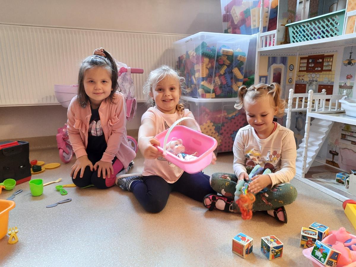 Na zdjęciu znajdują się 3 dziewczynki. Siedzą na podłodze, trzymają w rękach zabawki.