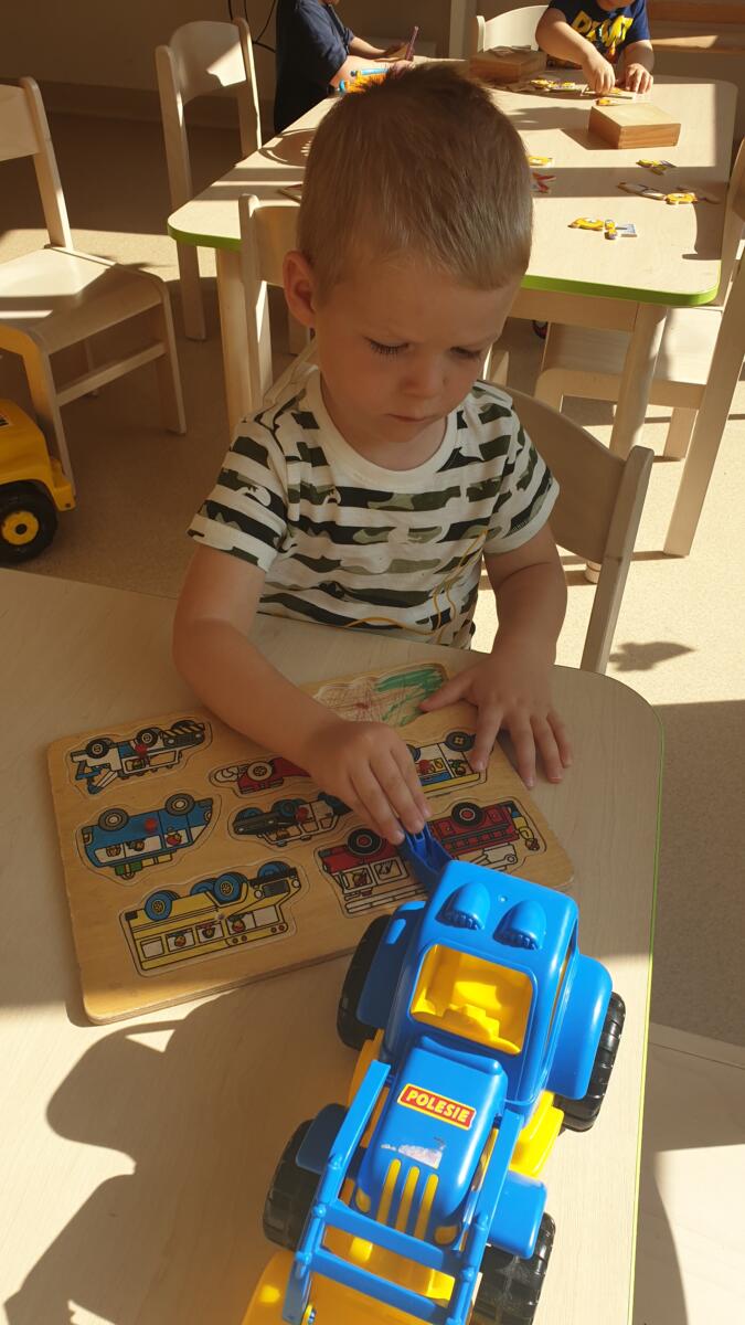 Chłopiec w koszulce w paski dopasowuje drewniane autka w odpowiednie miejsca układanki. Na jego twarzy maluje się skupienie.