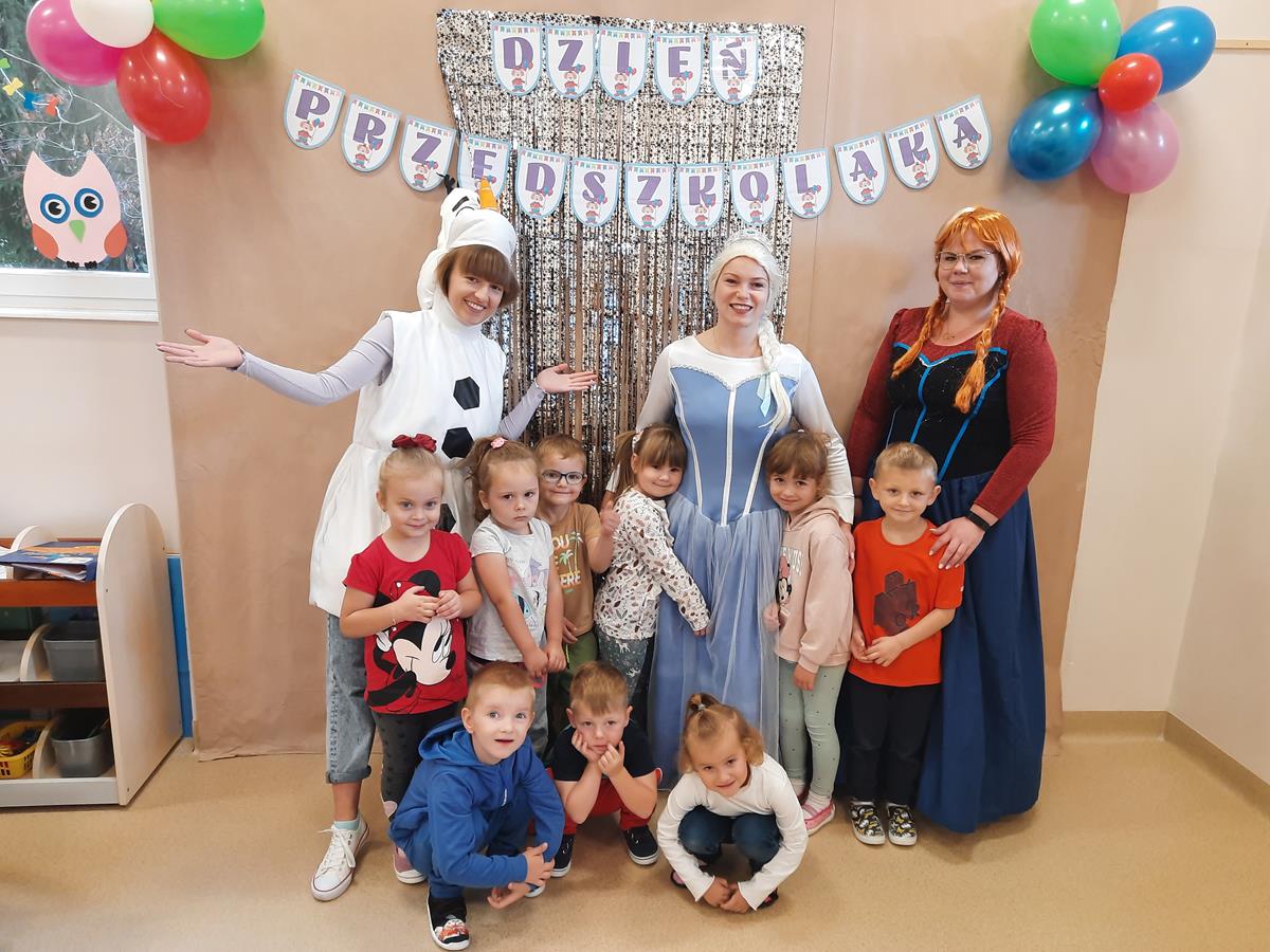 Grupa dzieci i osoby w bajkowych strojach: Elsy, Anny i Olafa pozują do zdjęcia. W tle na ścianie wisi napis "Dzień Przedszkolaka" oraz kolorowe balony.