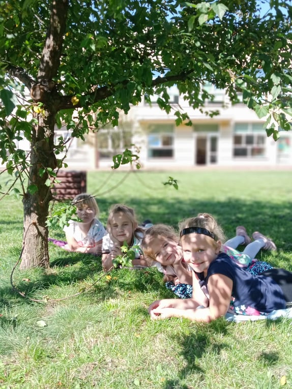 Na zdjęciu widać cztery dziewczynki, które leża pod drzewem w słoneczny dzień w ogrodzie przedszkolnym.
