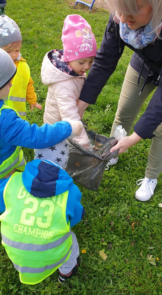 Przedszkolaki zbierają śmieci z trawnika i wrzucają do worka trzymanego przez panią.