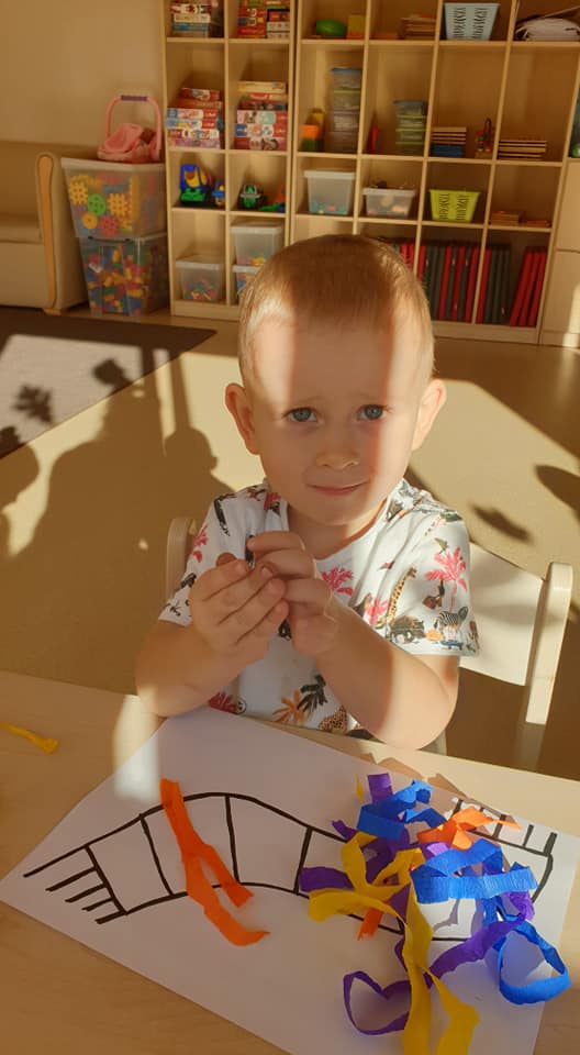 Zdjęcie przedstawia chłopca siedzącego przy stoliku, w miejscu pracy, który tworzy kuleczki z krepy.