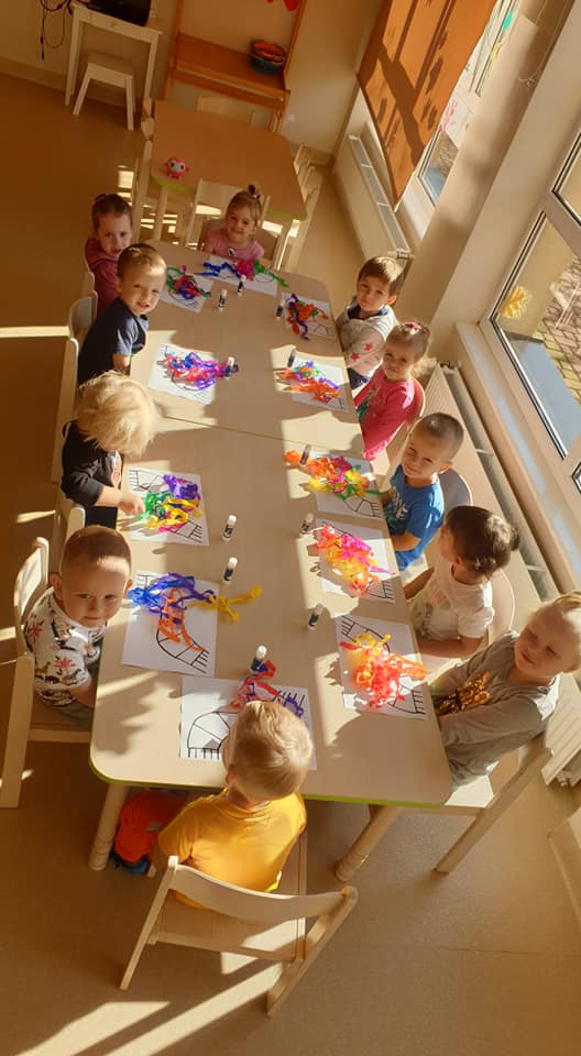 Dzieci z grupy "Niedźwiadki" siedzą pprzy stoliczku, na którym znajdują się kartki z wizerunkiem szalika, kolorowa krepa oraz kleje.
