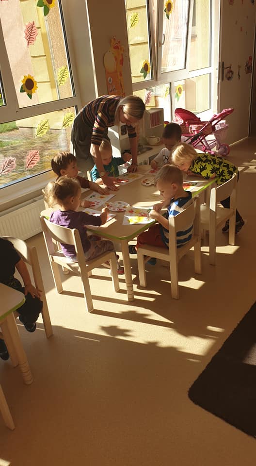 Przedszkolaki siedzące przy stoliku kontynuują malowanie farbkami kwiatków. Nauczyciel pomaga dzieciom w zadaniu.
