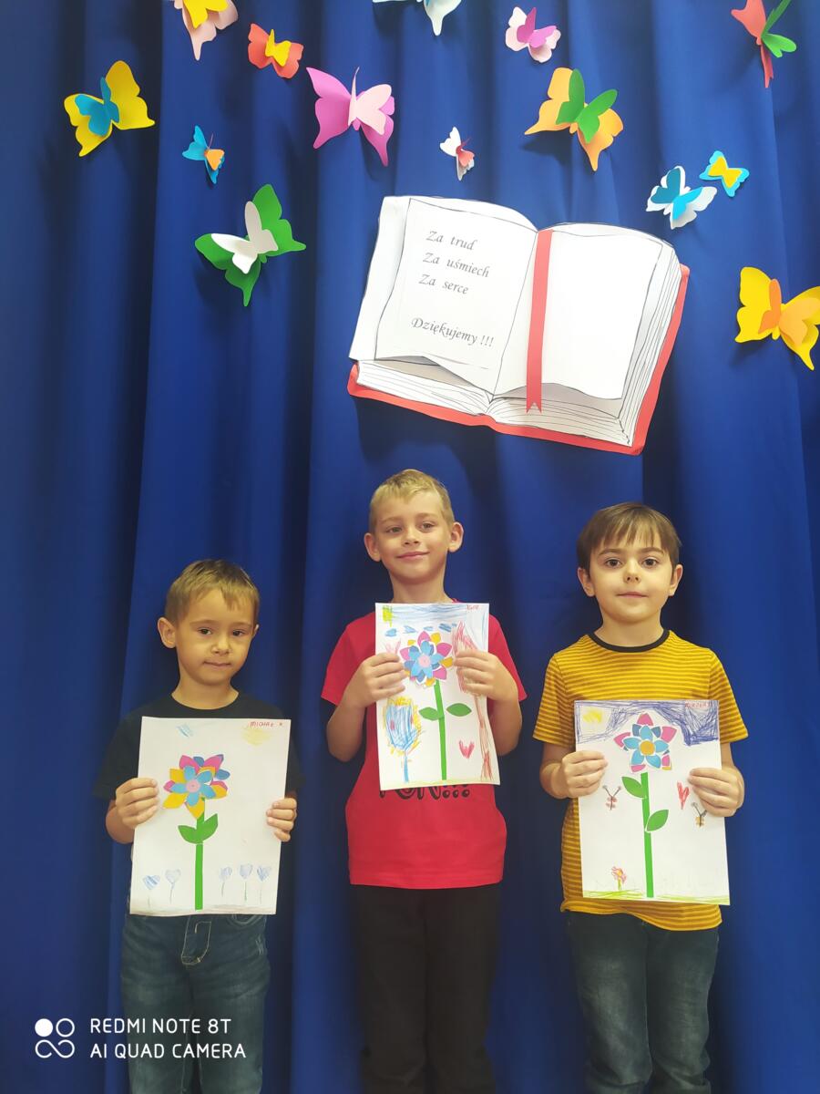 Na zdjęciu troje dzieci prezentuje wykonane przez siebie laurki - kwiatki dla nauczyciela.