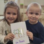 Na zdjęciu widzimy chłopca i dziewczynkę siedzących w sali przedszkolnej. Dzieci pozują do zdjęcia, trzymając w rączkach kartę obrazkową, ukazującą puzzle.