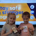 Na zdjęciu widzimy dwie dziewczynki stojące przed tablicą interaktywną. Dziewczynki pozują do zdjęcia, ukazują wykonane przez nie ksiązeczki, przedstawiające zasady zwalczania korona wirusa. Jest to książka w wersji mini. W tle na tablicy interaktywnej wyśwoetlony jest napis "stay safe& stronger together!'