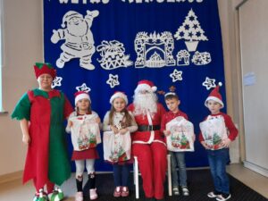 Zdjęcie przedstawia grupę dzieci w mikołajkowych czapkach wraz z Mikołajem i elfem.