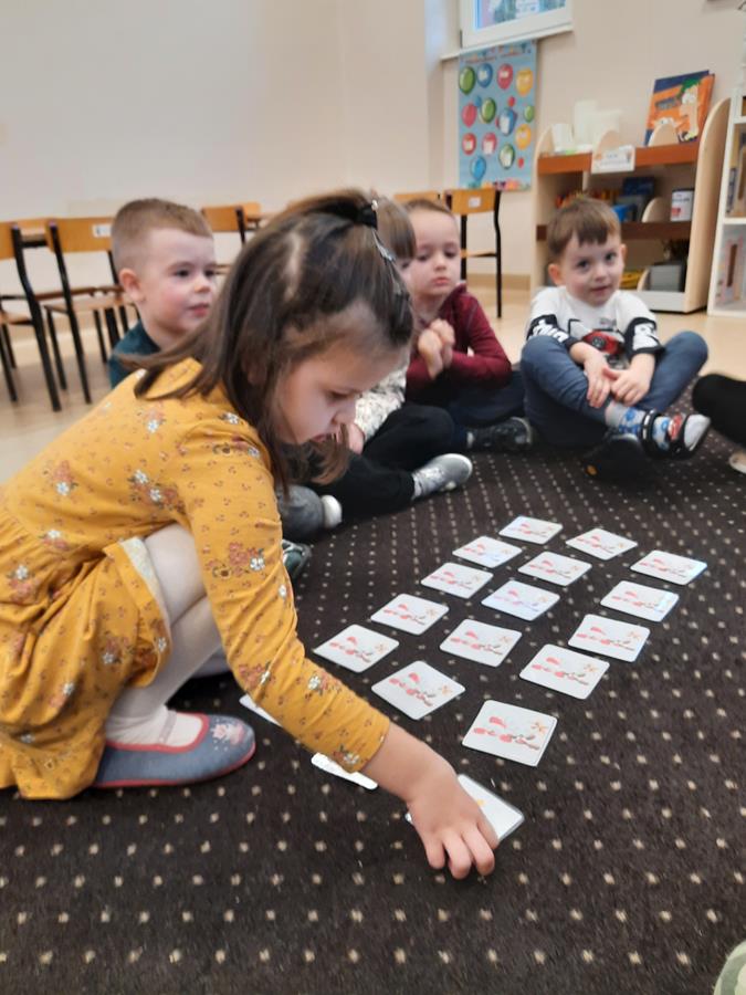 Dziewczynka dotyka małą kwadratową kartkę. Na dywanie koło niej siedzą inne dzieci, grają razem w grę memory.