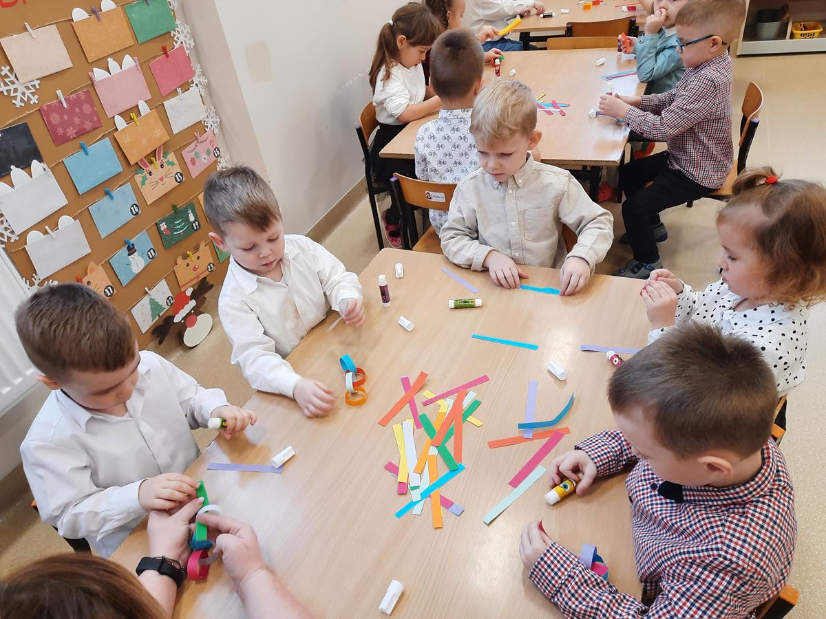 Kilkoro dzieci siedzi przy stoliku. Na środku stolika leżą paski kolorowego papieru i kleje. Dzieci z pasków robią kółka, które po połączeniu stworzą łańcuch.