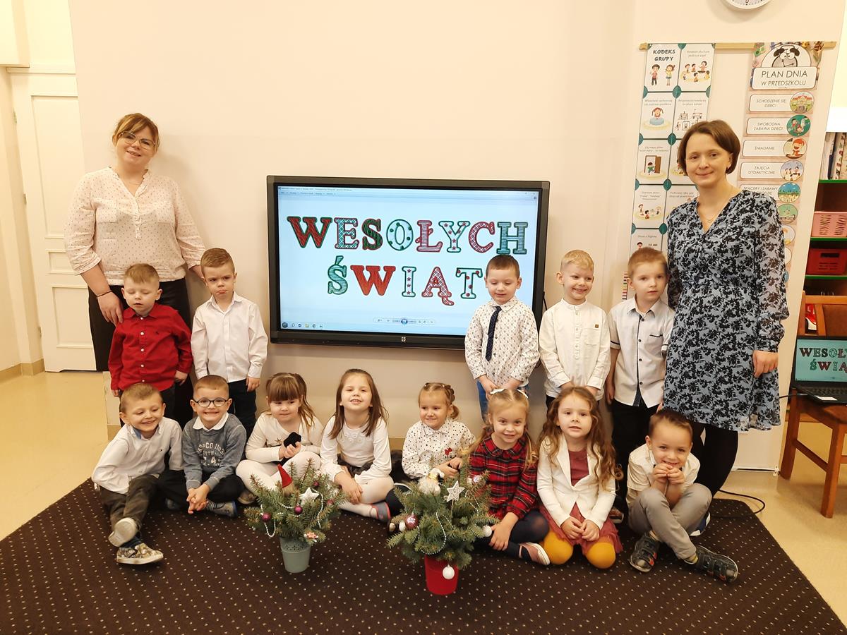 Dzieci wraz z paniami pozują do zdjęcia. W tle na tablicy multimedialnej wyświetlony jest napis "Wesołych Świąt".