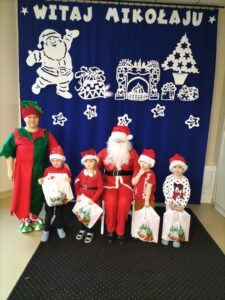 Dzieci w mikołajowych czapkach pozują do zdjęcia z Mikołajem i Elfem, w rękach trzymają otrzymane prezenty.