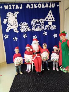 Dzieci w mikołajowych czapkach pozują do zdjęcia z Mikołajem i Elfem, w rękach trzymają otrzymane prezenty.