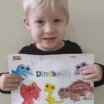 Na zdjęciu widzimy chłopca, który prezentuje swoją pracę. Pokolorowal na karcie pracy dinozaury..