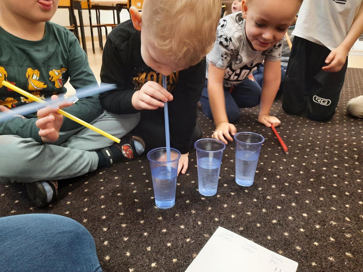 Chłopiec pije wodę przez słomkę. Na dywanie stoją 3 kubki plastikowe z wodą. Wokół chłopca siedzą pozostałe dzieci.