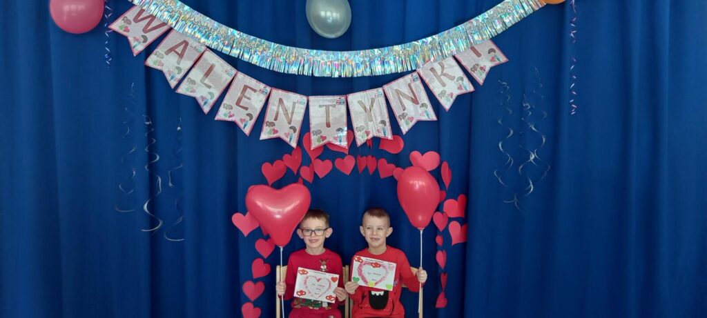 Zdjęcie przedstawia dwóch chłopców trzymających walentynki i czerwone sedruszka balony, w tle dekoracja z serduszek i napis "Walentynki".
