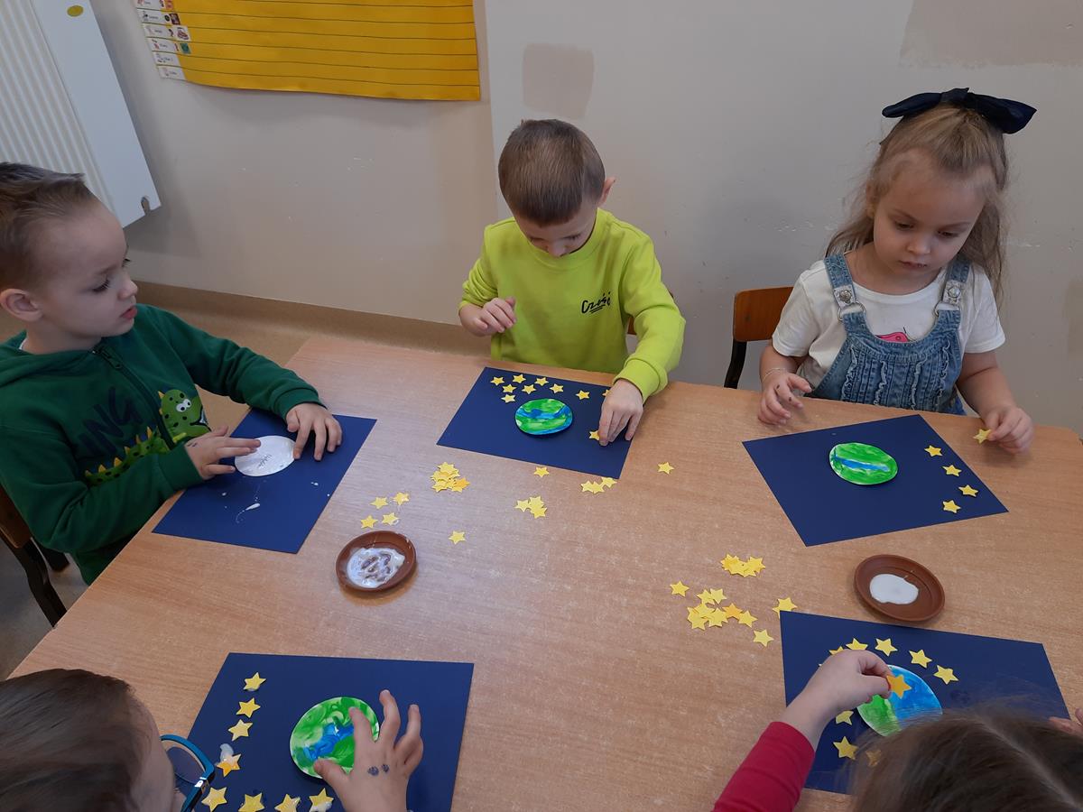 Dzieci siedzą przy stoliku i wykonują pracę plastyczną "Ziemia wśród gwiazd" naklejając gwiazdki z papieru wokół planety.
