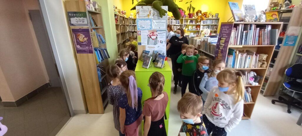 Grupa Delfinki zwiedza bibliotekę, dzieci poruszaja się między regałami z książkami.