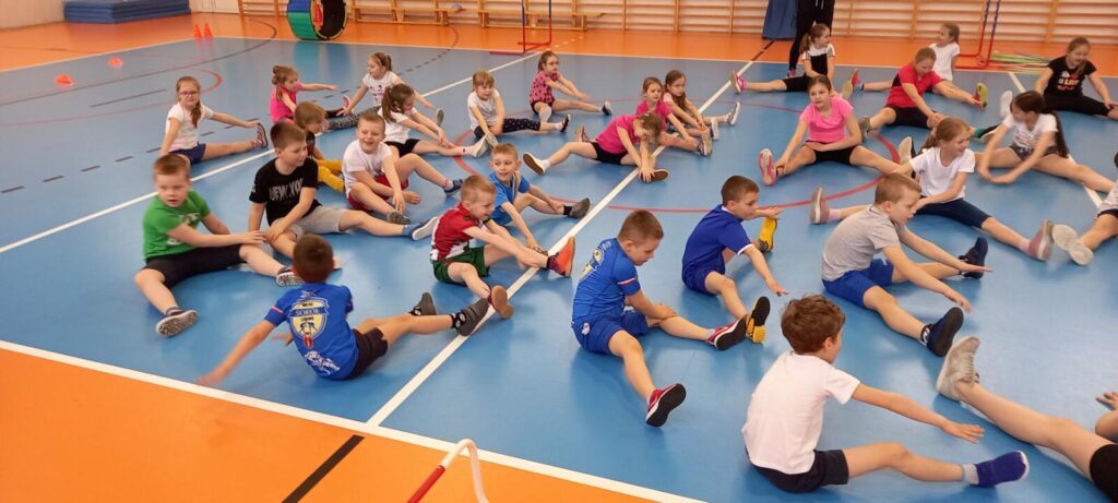 Zdjęcie przedstawia dzieci podczas zajęć sportowych w sali gimnastycznej.