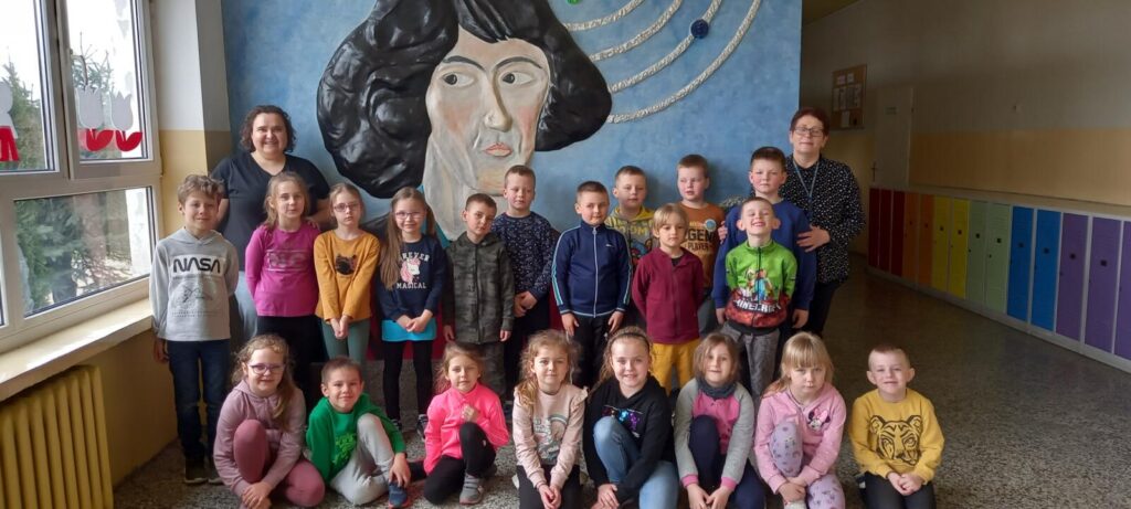 Grupa Delfinki pozuje do zdjęcia na korytarzu szkoły - w tle dzieci namalowana postać Mikołaja Kopernika patrona szkoły