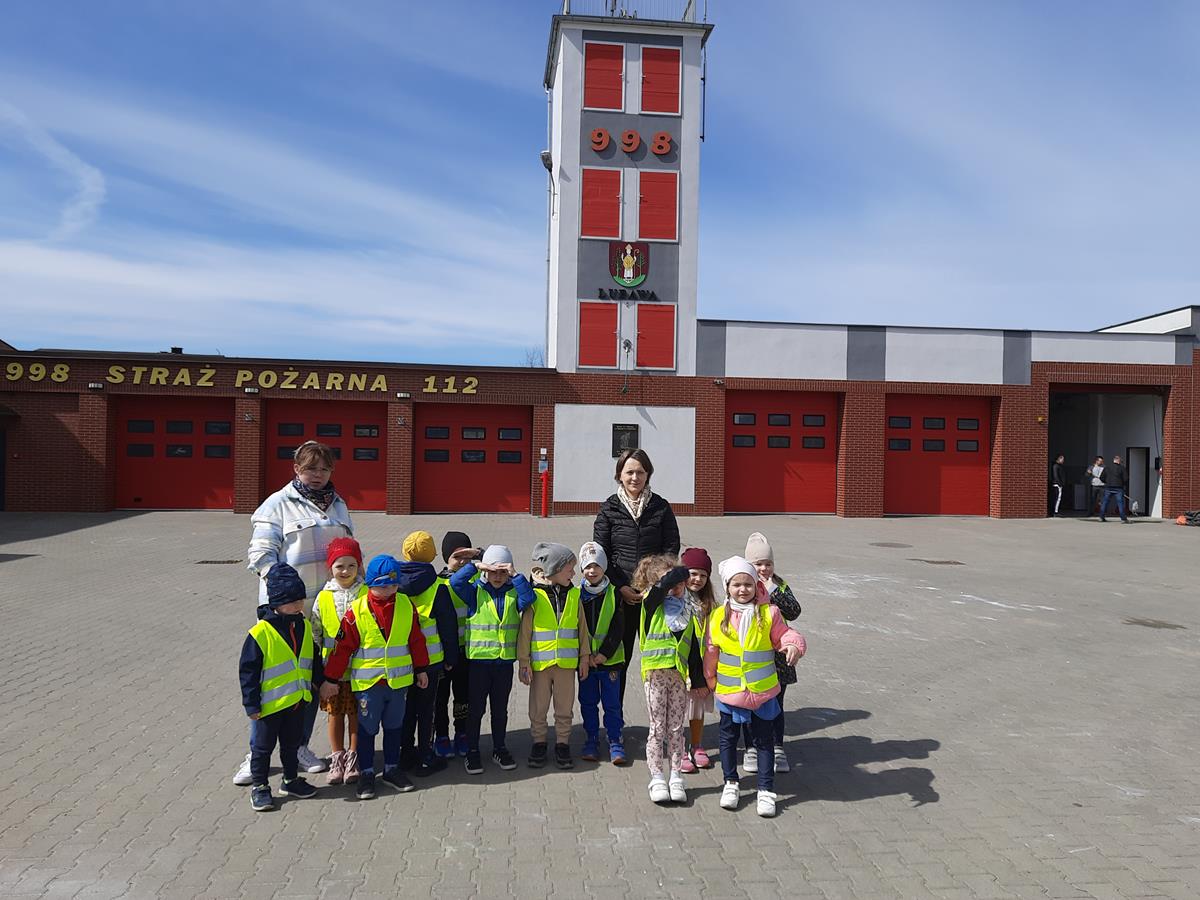 Dzieci wraz z nauczycielami pozują do zdjęcia, w tle znajduje się budynek Ochotniczej Straży Pożarnej w Lubawie.