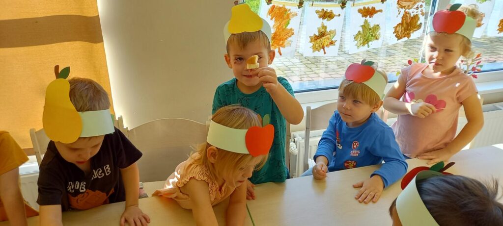 Kilkoro dzieci w kolorowych - owocowych opaskach na głowie za pomocą wykałaczek wykonują owocowe szaszłyki.
