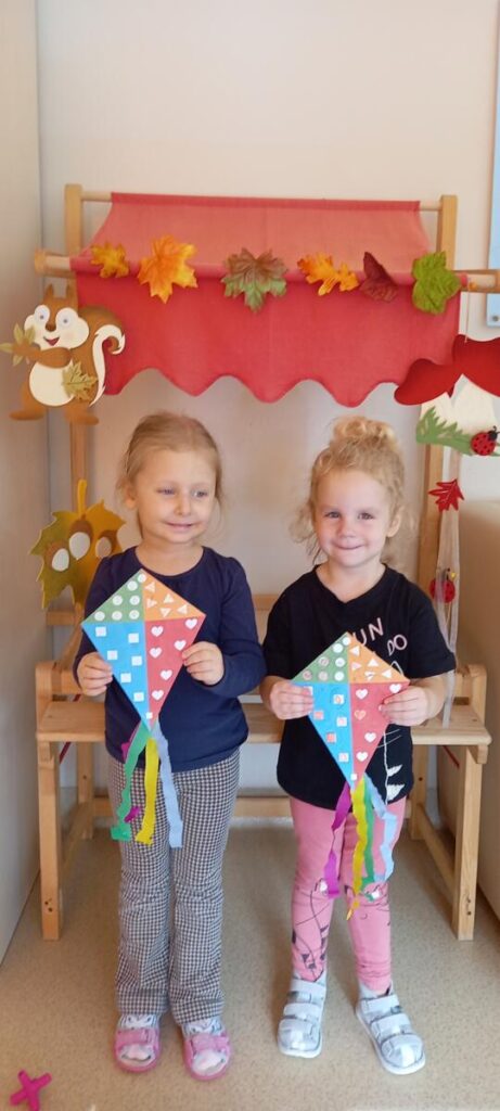 Dwie dziewczynki stojąc na tle jesiennej dekoracji- prezentują swoje wykonane papierowe latawce.