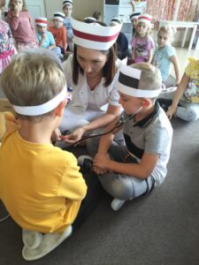 Zdjęcie przedstawia dzieci, którym pani pielęgniarka pokazuje w jaki sposób działa stetoskop. Jeden chłopiec osłuchuje drugiego.