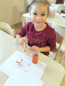Zdjęcie przedstawia dziewczynkę,ktoś maluje farbami na pomarańczowo rolkę po papierze toaletowym.
