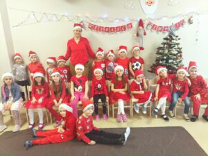Zdjęcie przedstawia dzieci wraz z wychowawcą,którzy pozują do zdjęcia na świątecznym tle. Wszyscy mają ubrane czerwone bluzki i mikołajowe czapki.