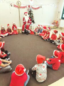 Zdjęcie przedstawia dzieci siedzące w kole wraz z Mikołajem. Dzieci w mikołajkowych czapkach śpiewają piosenkę dla Mikołaja.
