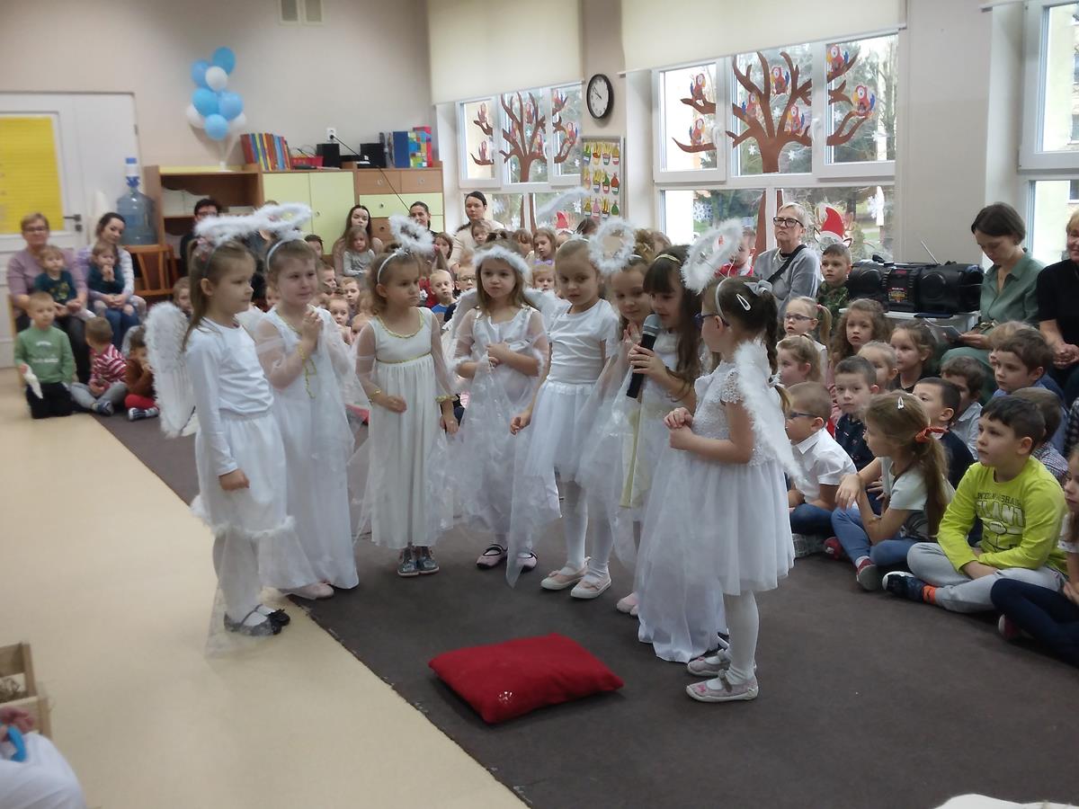 Dziewczynki w strojach aniołków mówią swoje kwestia stojąc przed stajenką. W tle siedzi duża liczba dzieci oglądający przedstawienie.