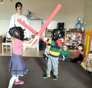 Zdjęcie przedstawia dwoje dzieci,które toczą "walkę" na piankowe wałki. Na głowie mają specjalne kaski ochronne.